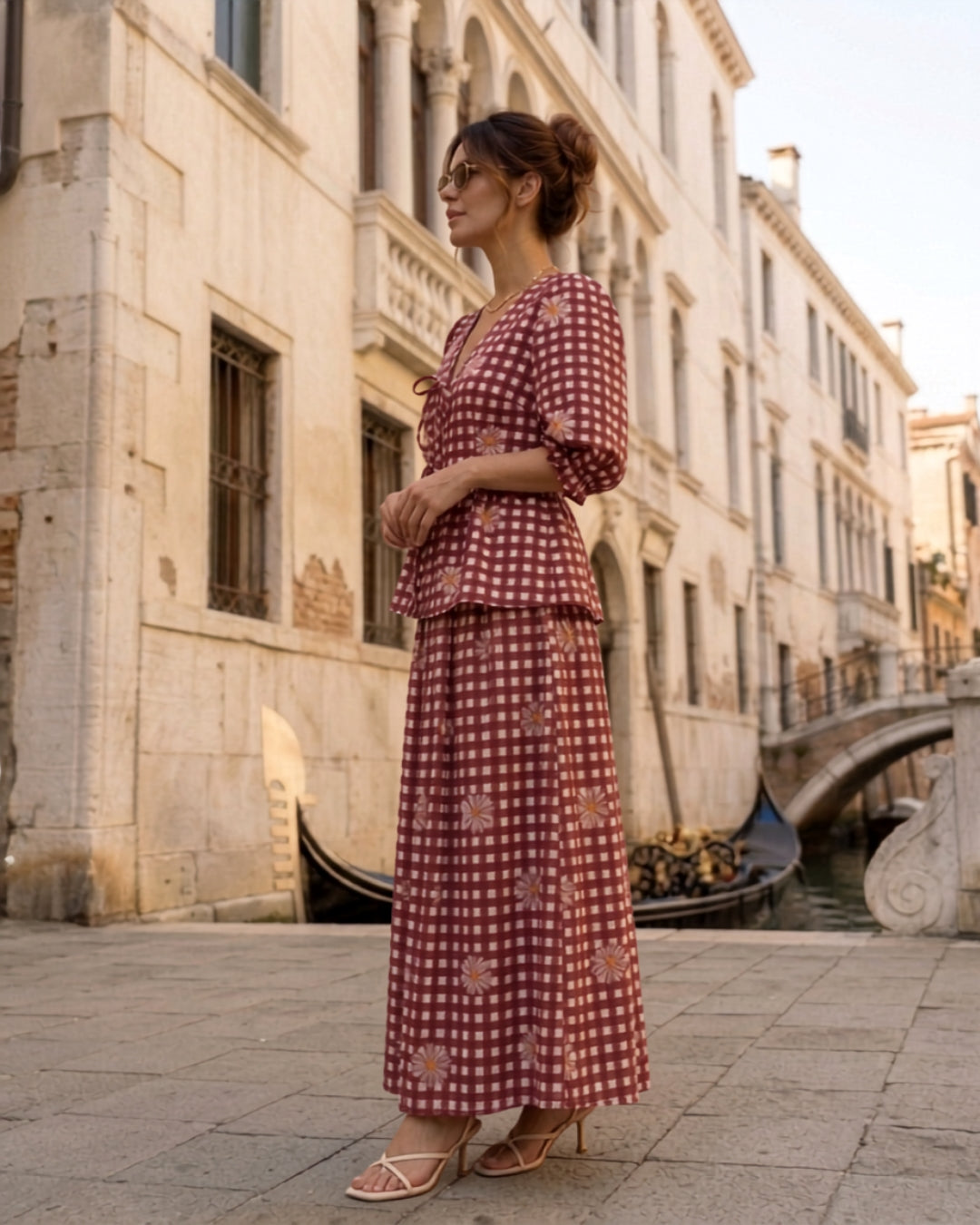 A woman wearing a red and white checkered two-piece set with a peplum waist and matching skirt, standing on a stone walkway in front of a canal and historic building.