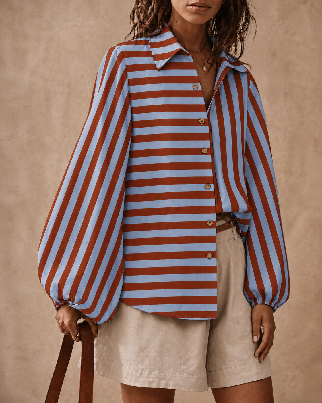 A woman wearing a long-sleeve button-front shirt with vertical rust red and light blue stripes, paired with light beige shorts, standing against a warm beige textured backdrop.