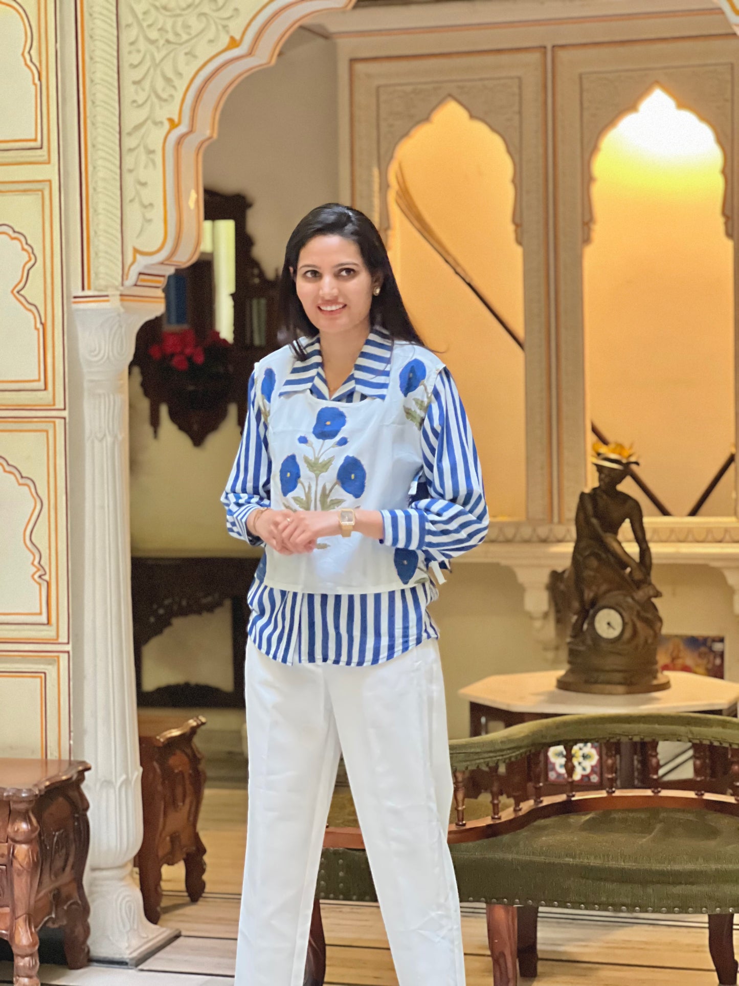 Woman in a blue and white floral blouse with white pants standing in an ornate room.