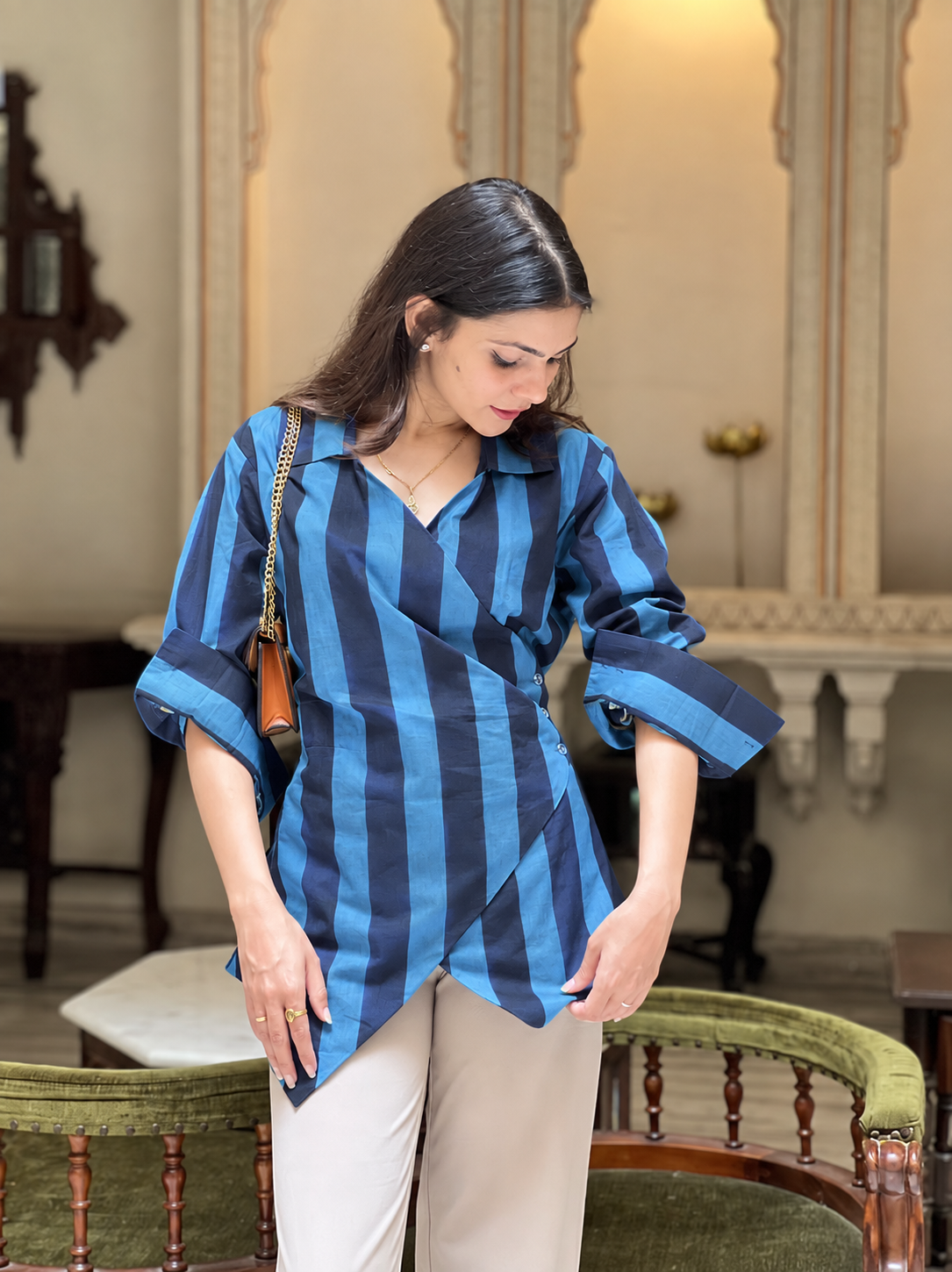 A woman wearing a blue and navy vertically striped wrap shirt with beige trousers, standing in an elegant indoor setting with a mirror and wooden furniture in the background.