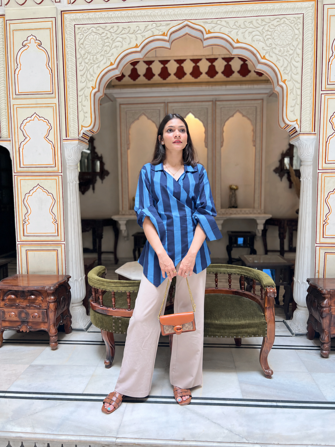 A woman stands in front of an ornate architectural archway, wearing a blue and navy striped button-up shirt, beige wide-leg trousers, and holding a small orange handbag.