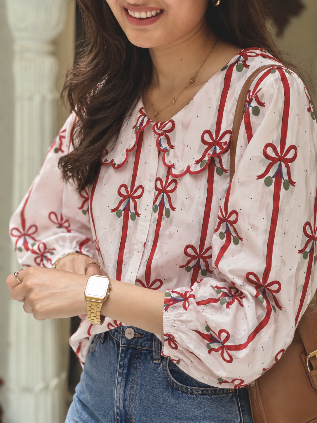 A woman wearing a light pink blouse with red bow and ribbon patterns, paired with blue jeans and a gold watch.
