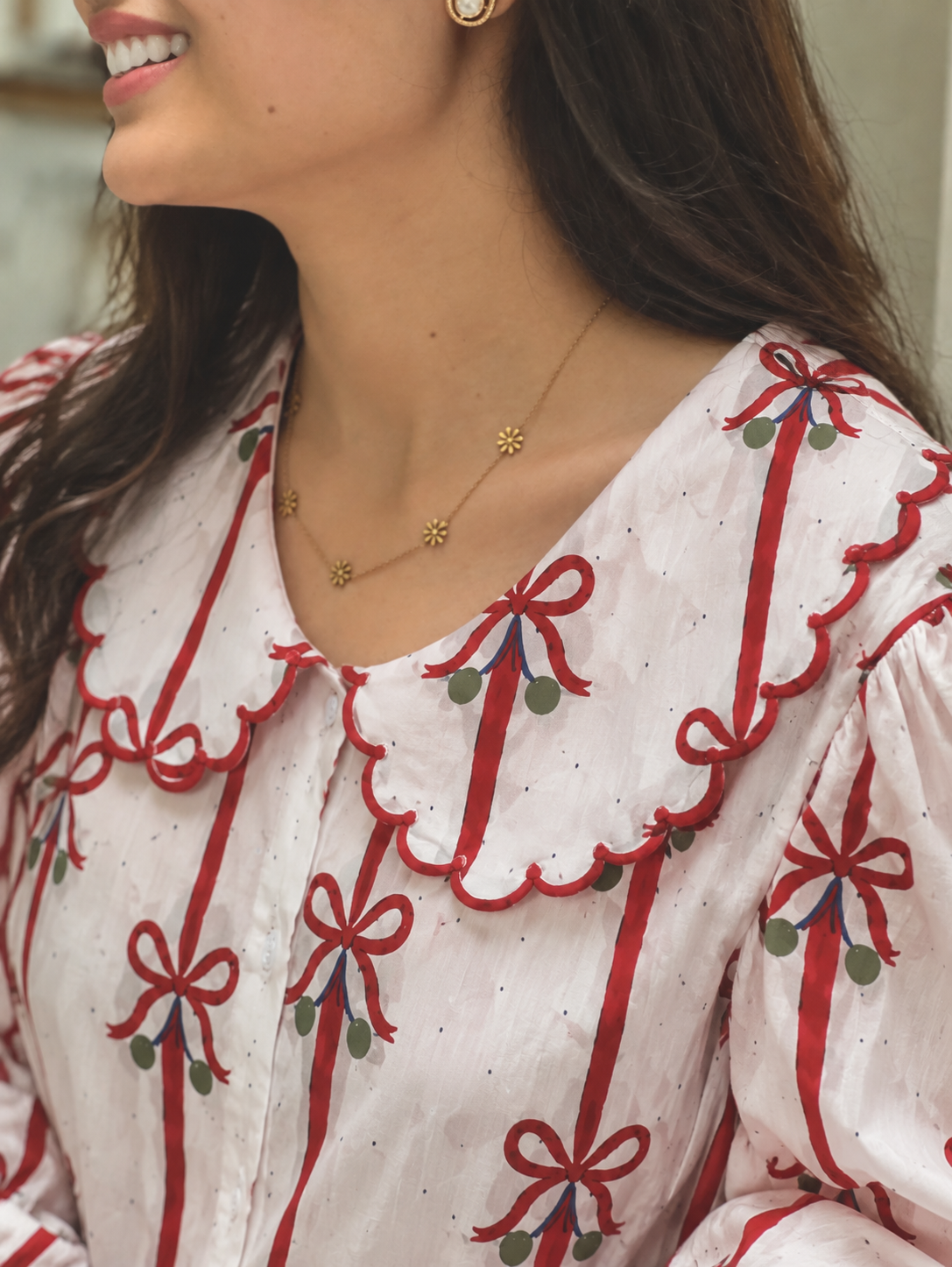 A close-up of a woman wearing a white blouse with red ribbon and green berry embroidery, a scalloped ruffled collar, and gold floral chain necklaces.
