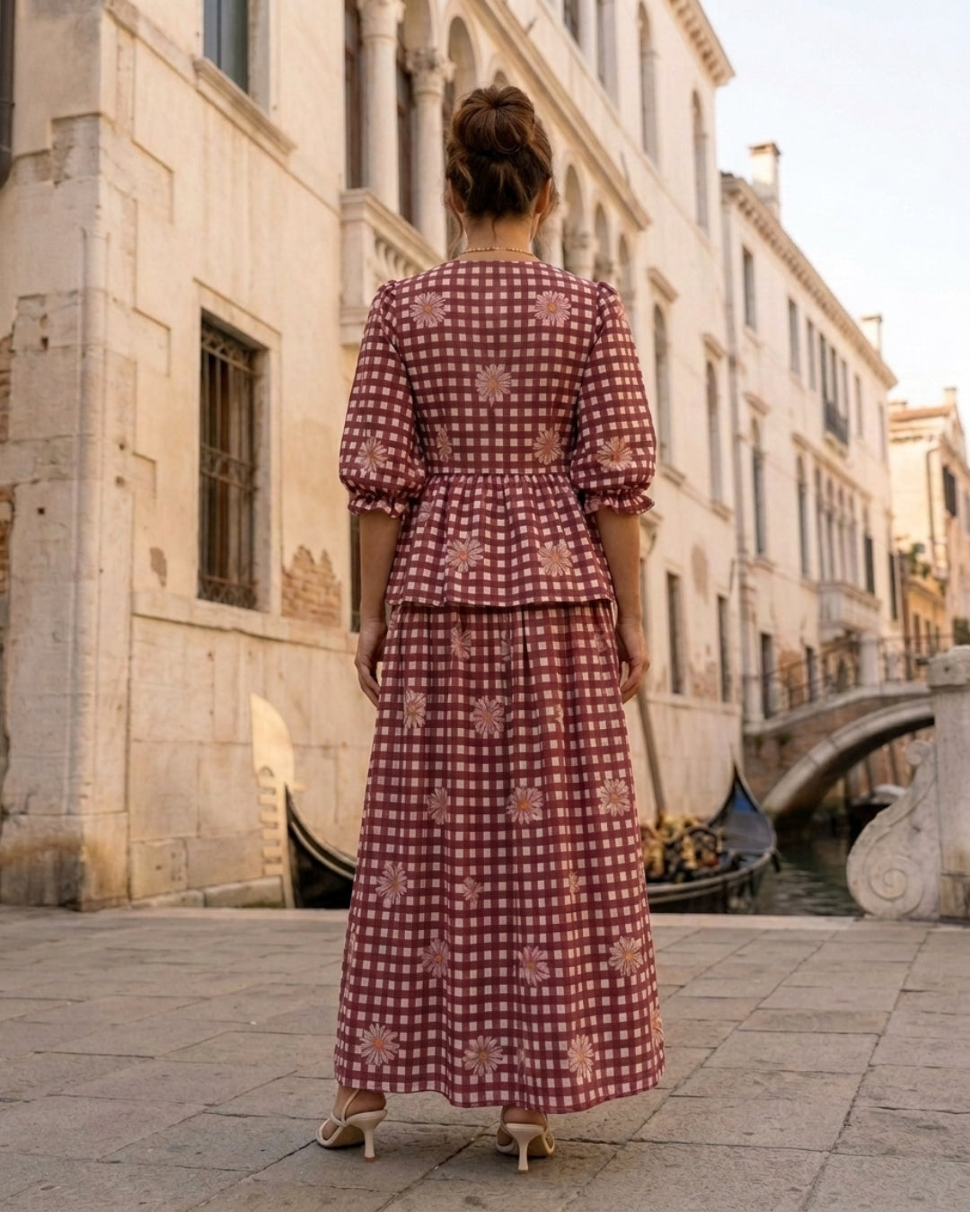 A woman viewed from behind wearing a long-sleeved red and white gingham dress with a peplum waist, standing on a stone walkway in front of historic Venetian buildings and a canal.