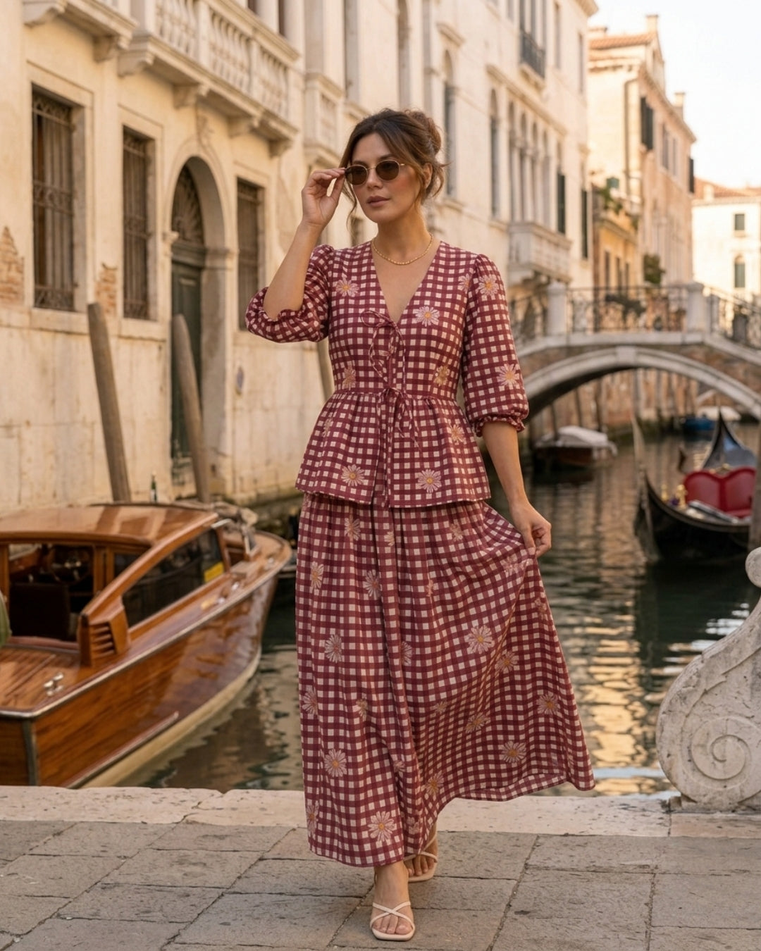 A woman wearing a red and white gingham two-piece set with a floral print stands on a stone bridge in Venice, with a canal, boats, and historic buildings in the background.