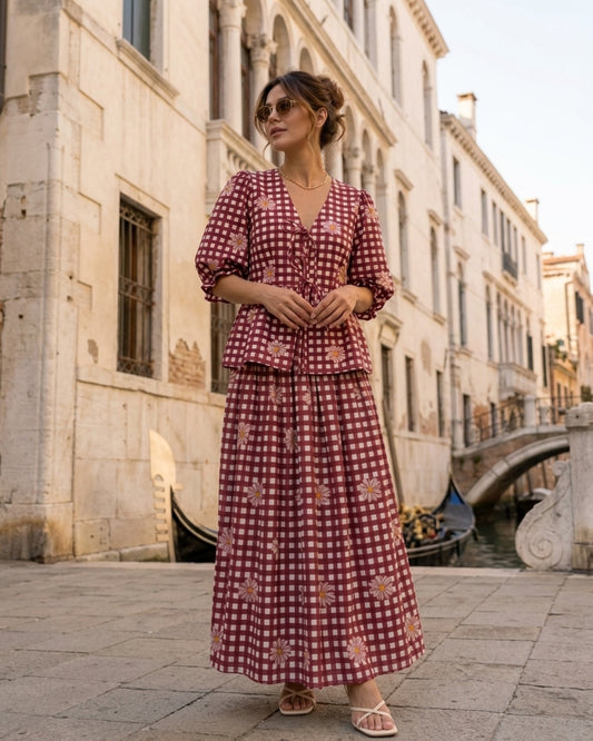 A woman wearing a red and white gingham two-piece dress stands on a stone walkway in a canal-side city setting with historic buildings and a bridge in the background.