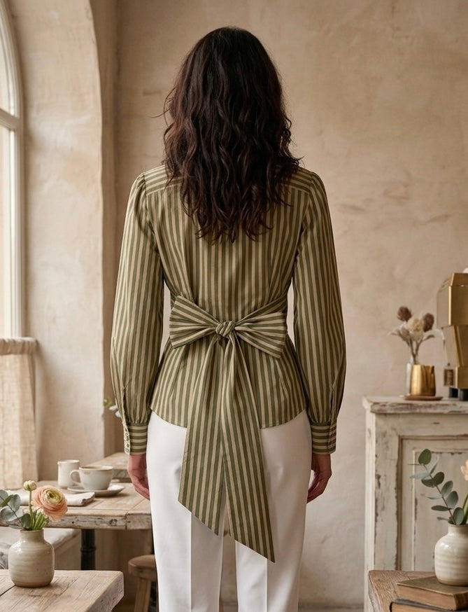 Woman wearing a green striped shirt with a tie waist in a room with a window and furniture.