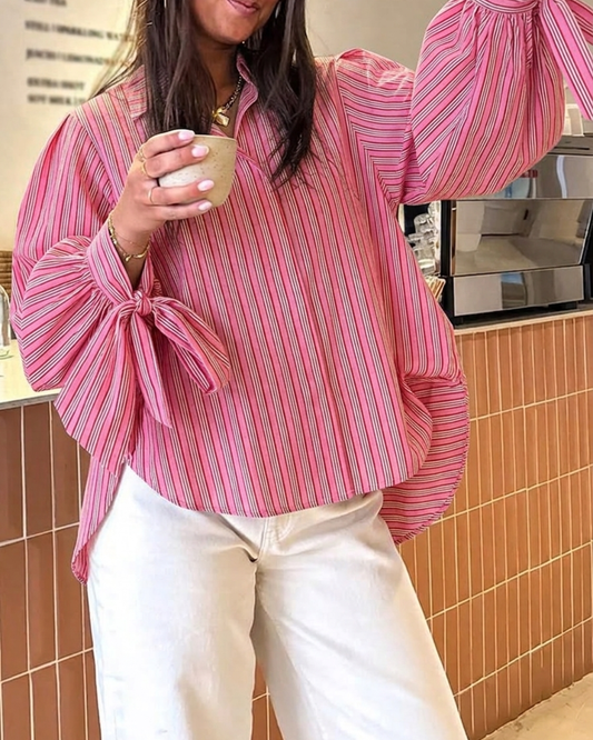 A woman wearing a pink and red vertically striped blouse with dramatic balloon sleeves and side tie details, paired with cream-colored pants, holding a small cup in a cafe setting.