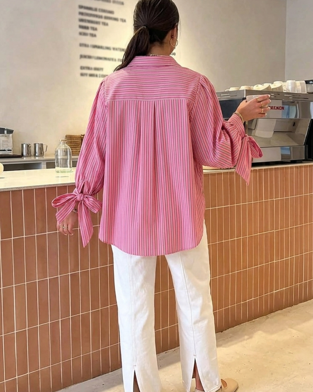 A woman viewed from behind wearing a pink and white vertically striped button-up shirt with tie details on the sleeves, paired with white pants, standing in a cafe setting.