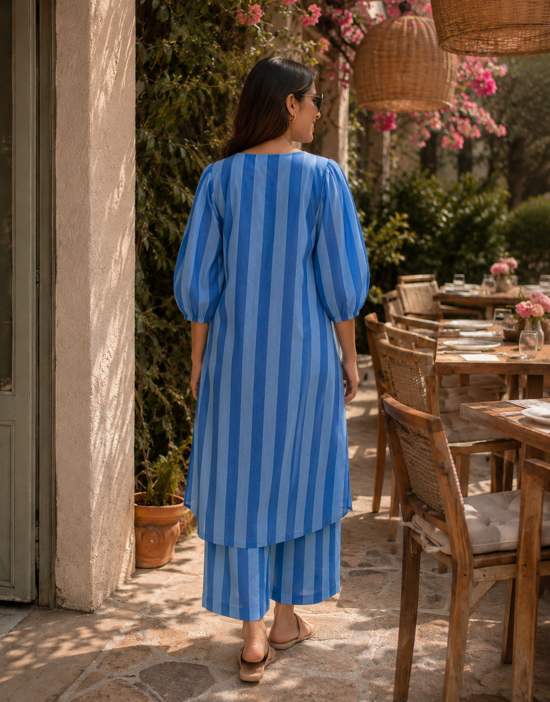 A woman viewed from behind wearing a blue and light blue vertically striped kurta and matching pants in an outdoor patio setting.