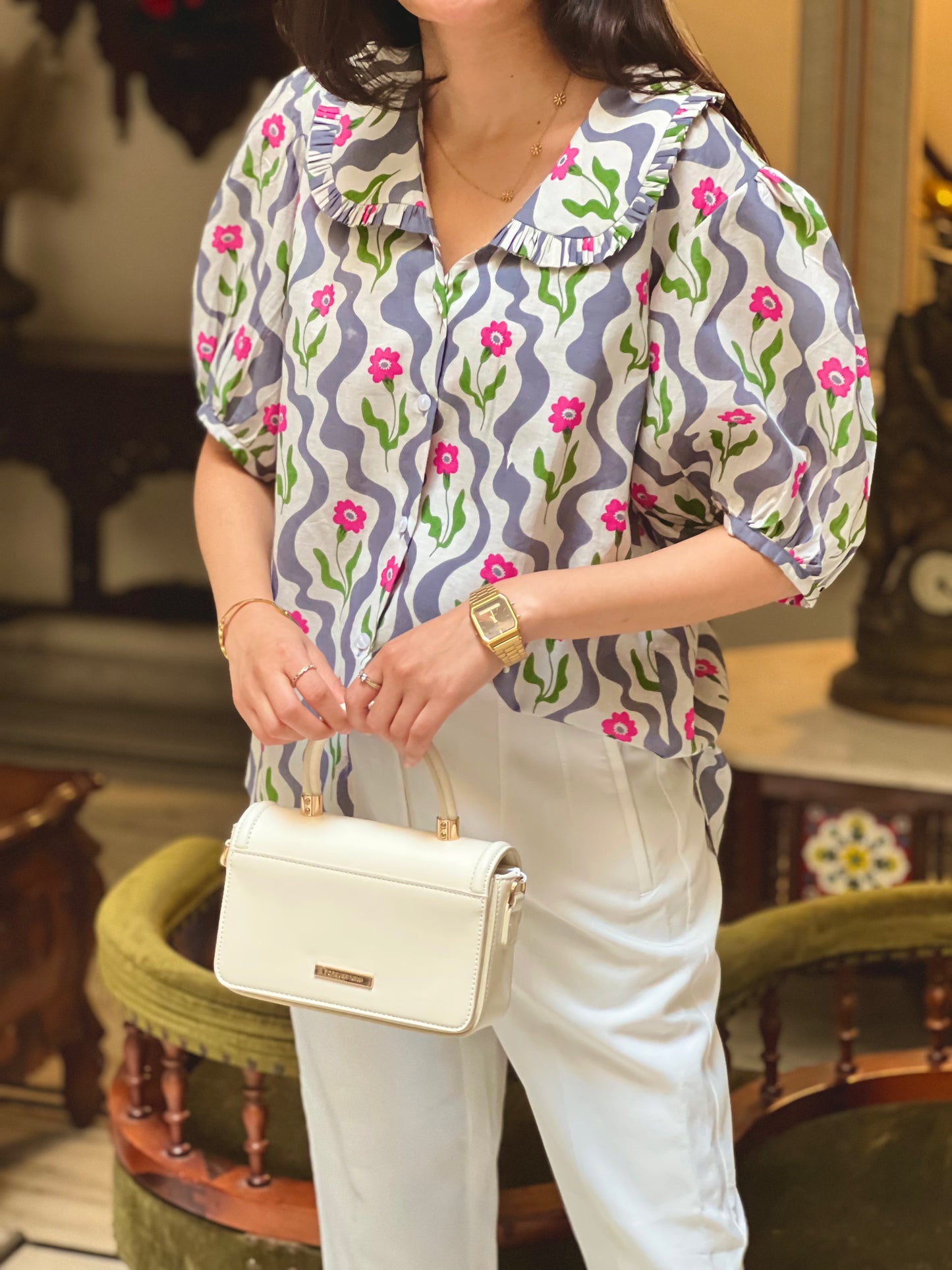 Woman holding a white handbag wearing a patterned blouse and white pants in an indoor setting.