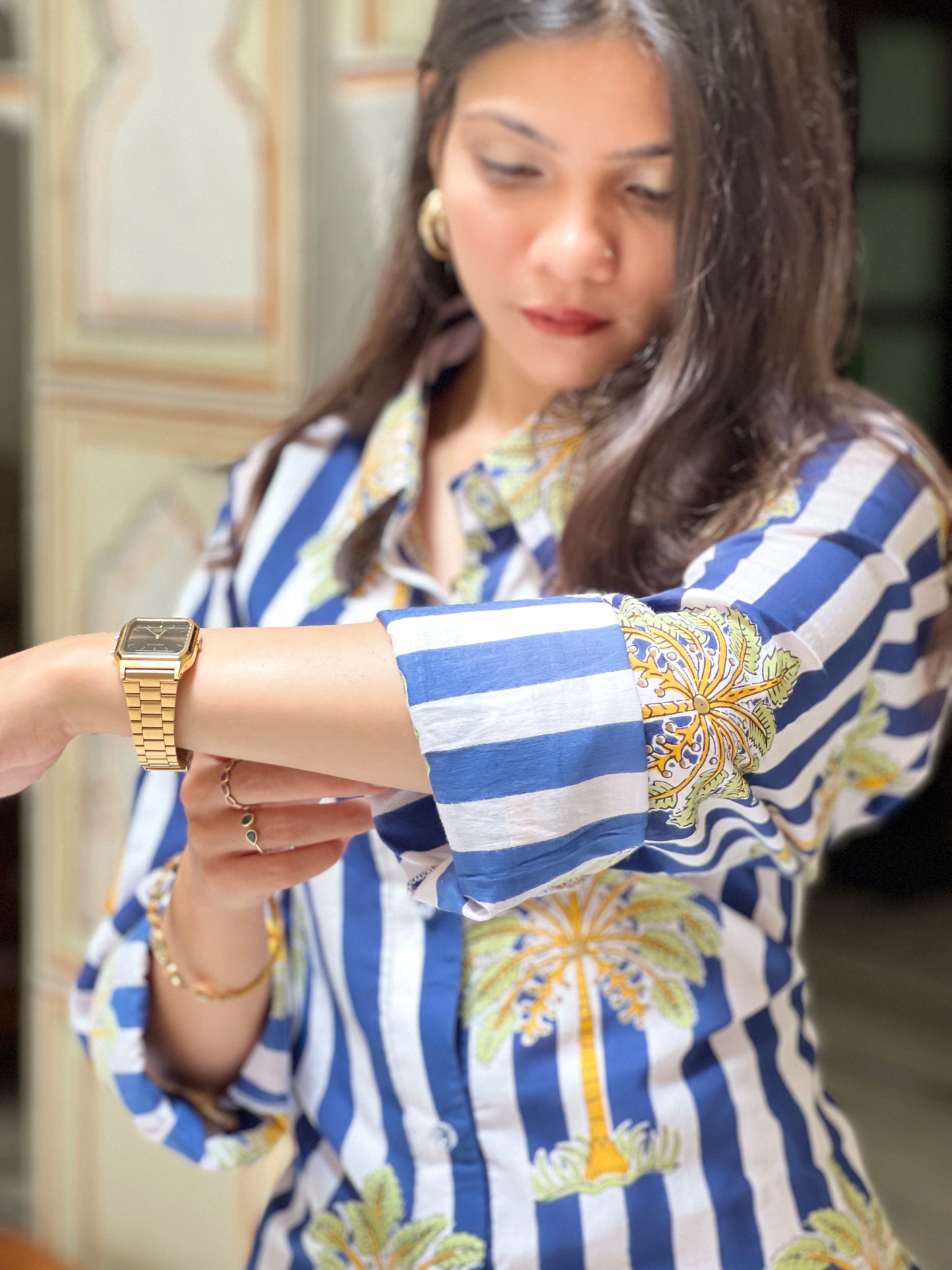 Woman wearing a blue and white striped shirt with floral patterns, looking at her gold watch.