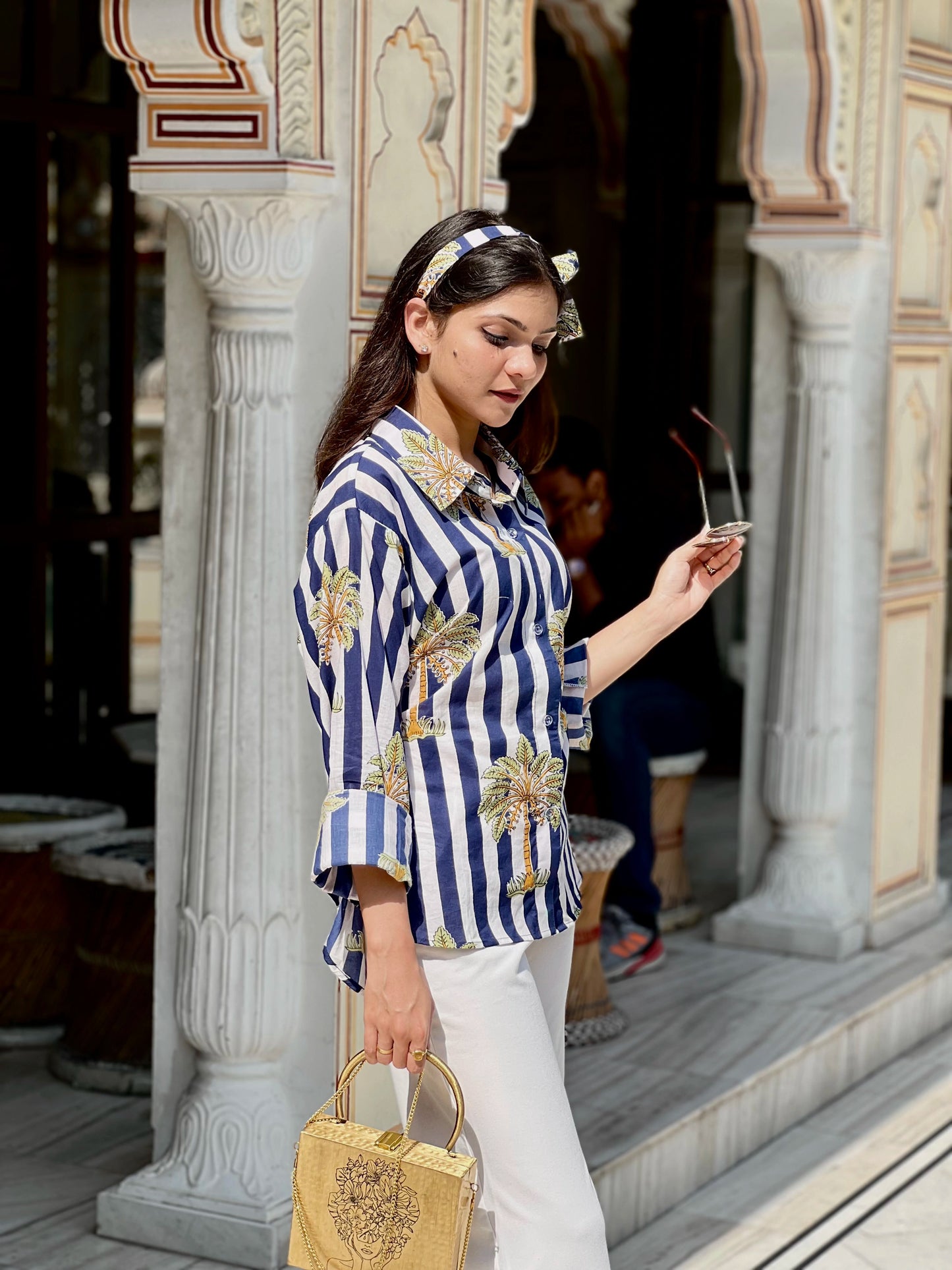 Woman in a blue and white striped shirt with floral patterns, holding a beige handbag, standing in front of decorative columns.