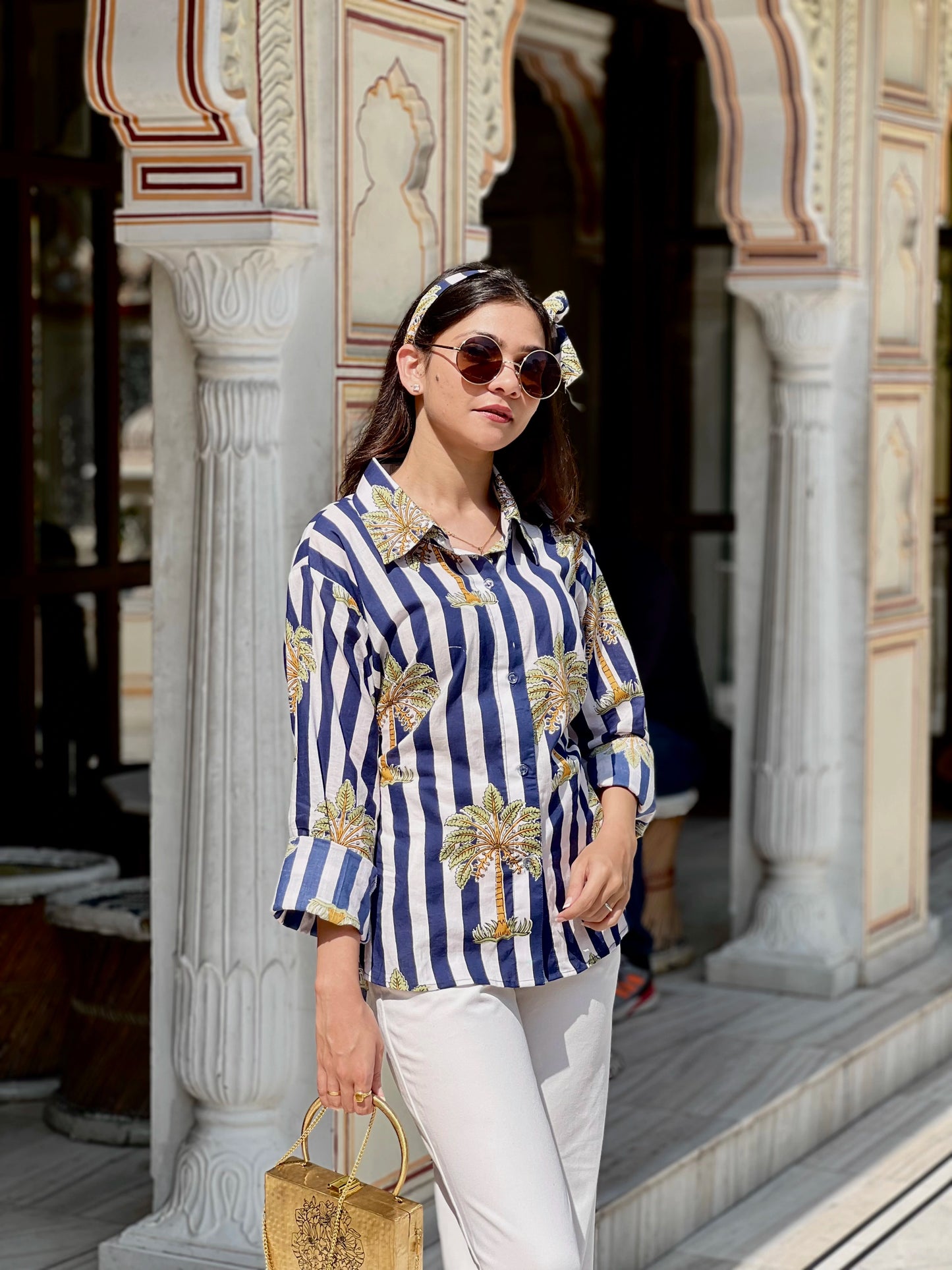 Woman wearing a blue and white striped shirt with palm tree patterns, standing in front of decorative columns.