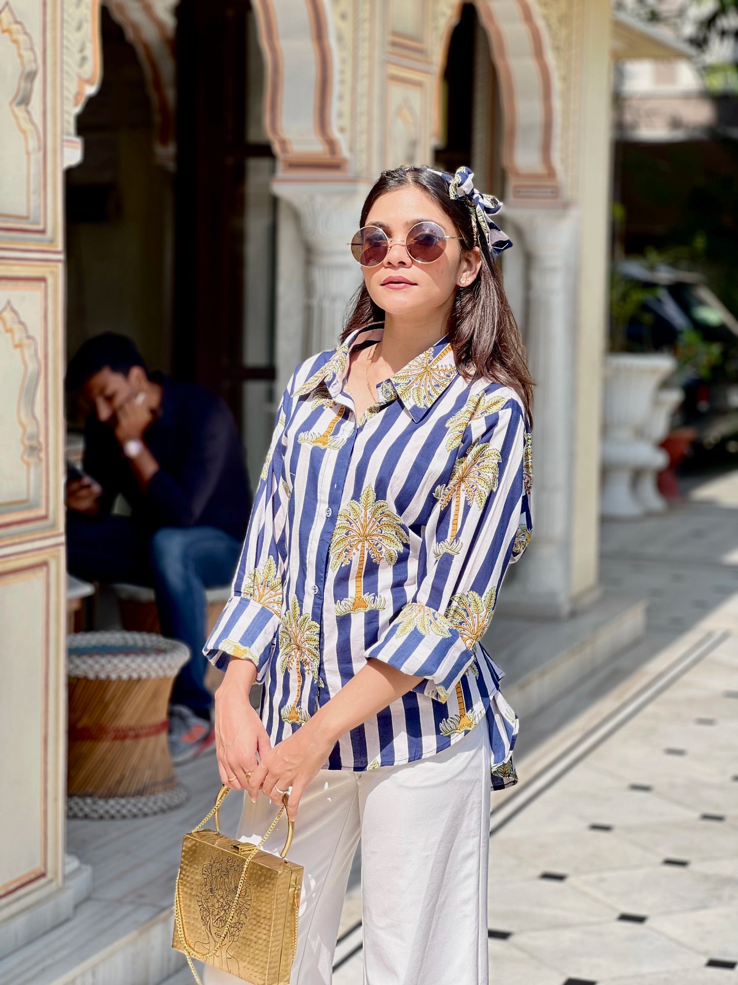 Woman in a blue and white striped shirt with palm tree design, holding a straw bag, standing in front of an architectural building.