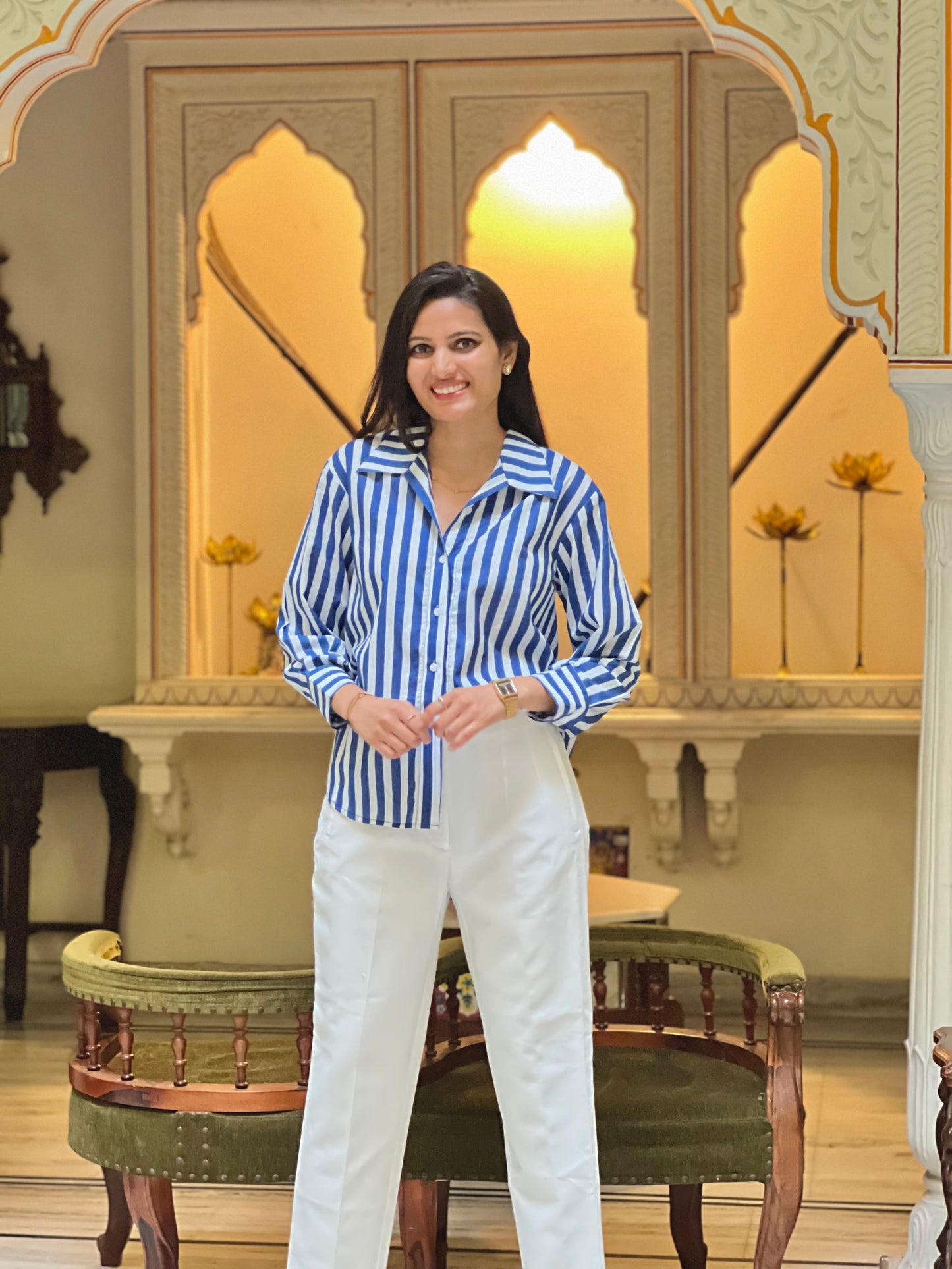 Woman in a blue and white striped shirt and white pants standing in an ornate room.