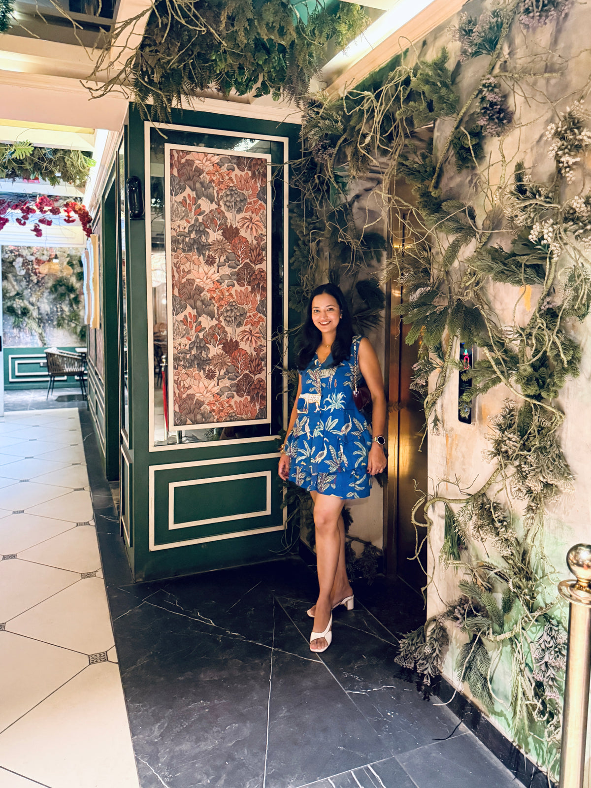 A woman wearing a blue floral sleeveless dress stands in an indoor hallway next to a green decorative cabinet, with a wall covered in greenery on the right.