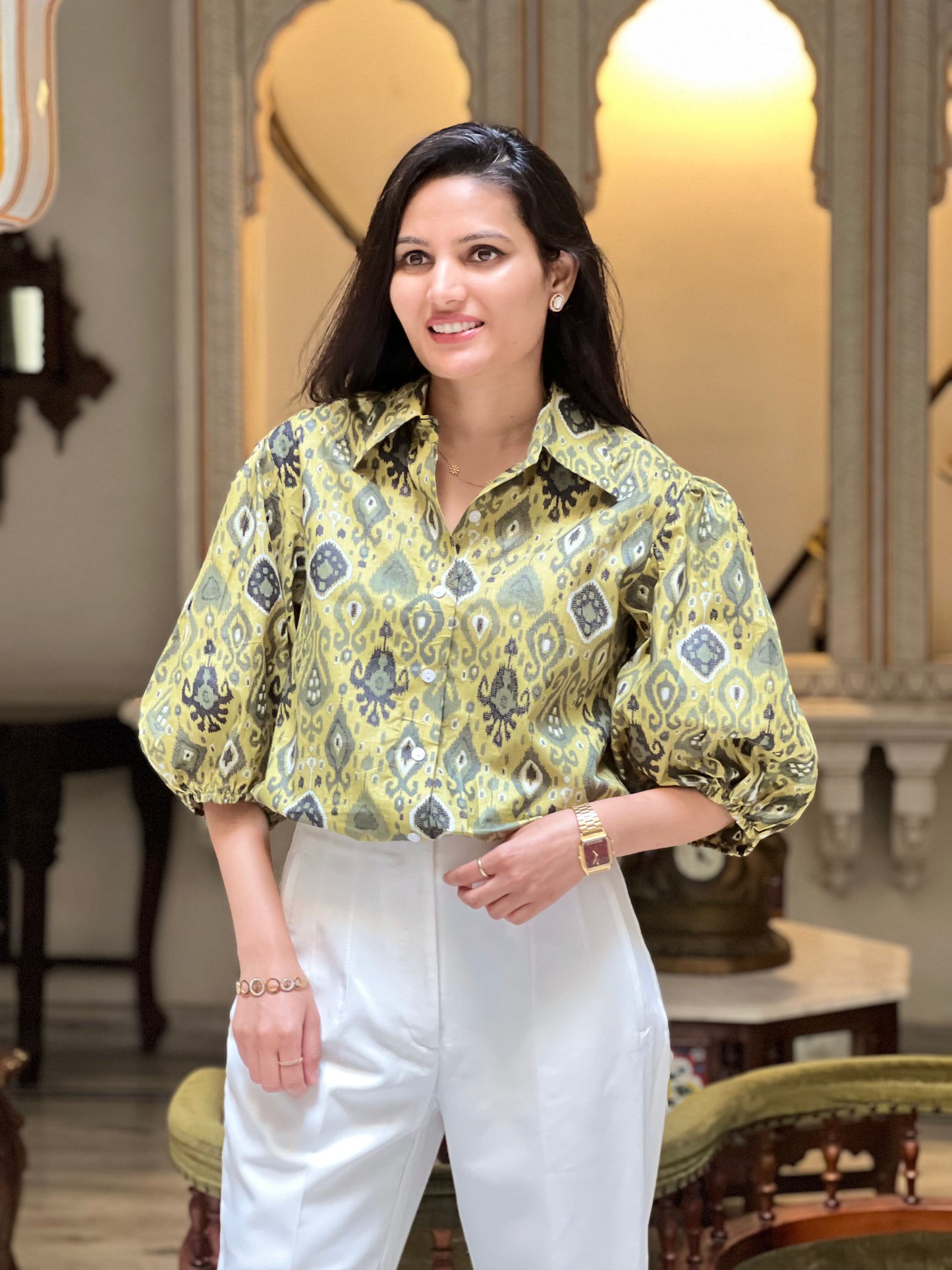 Woman wearing a patterned yellow blouse and white pants in an indoor setting.