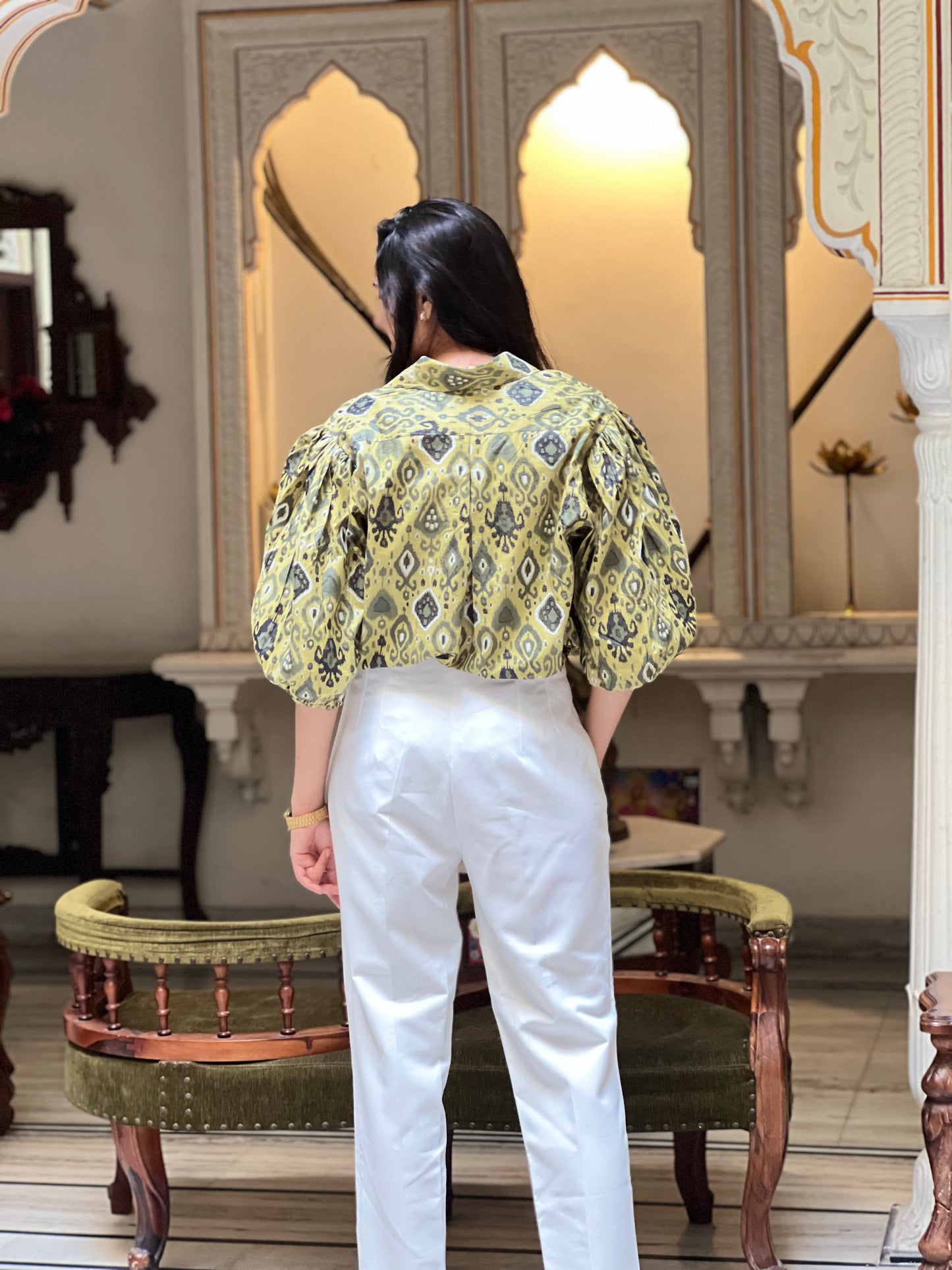 Person wearing a patterned top and white pants in an ornate room.