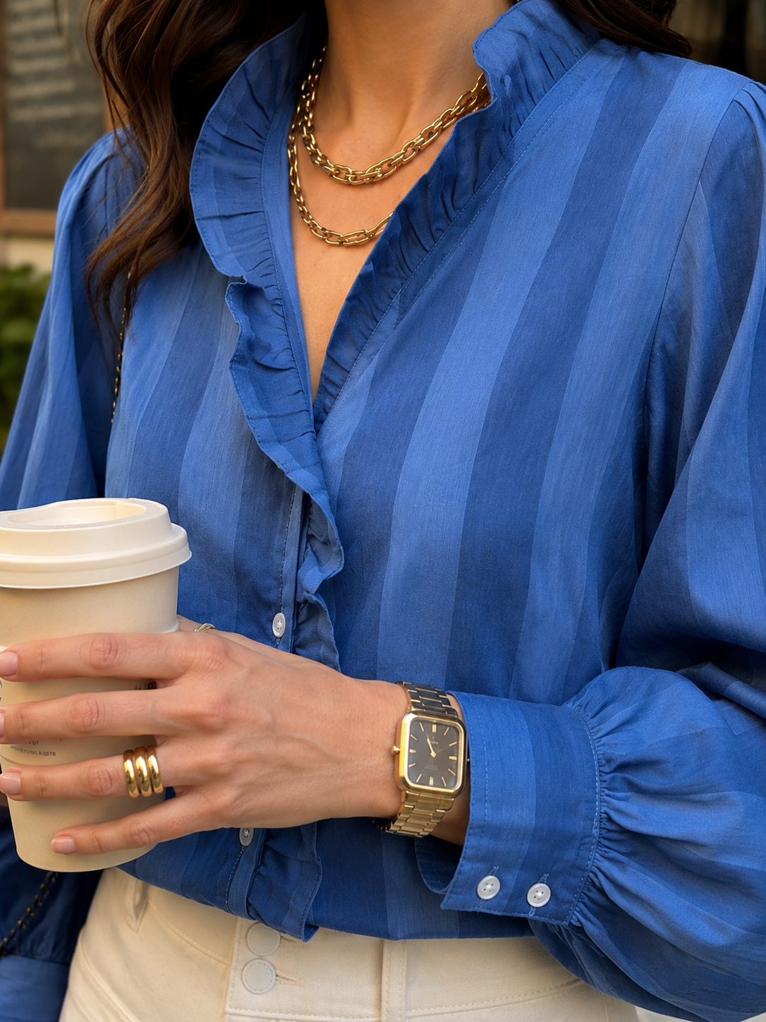 Person wearing a blue blouse, holding a coffee cup, with gold jewelry.