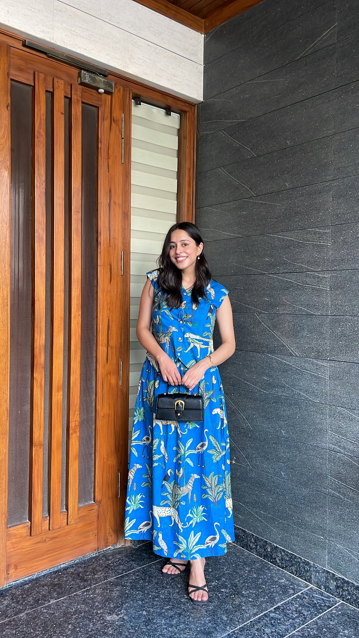 Woman in a blue dress standing in front of a wooden door and gray wall.