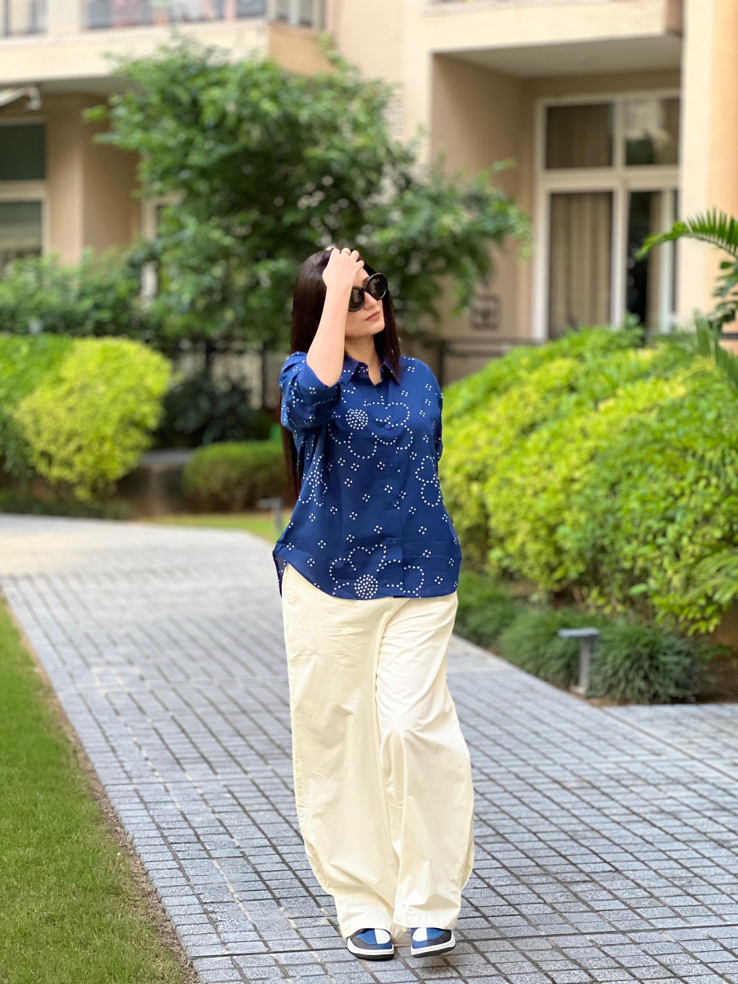 Woman in a blue patterned shirt and beige pants walking on a paved path with greenery in the background