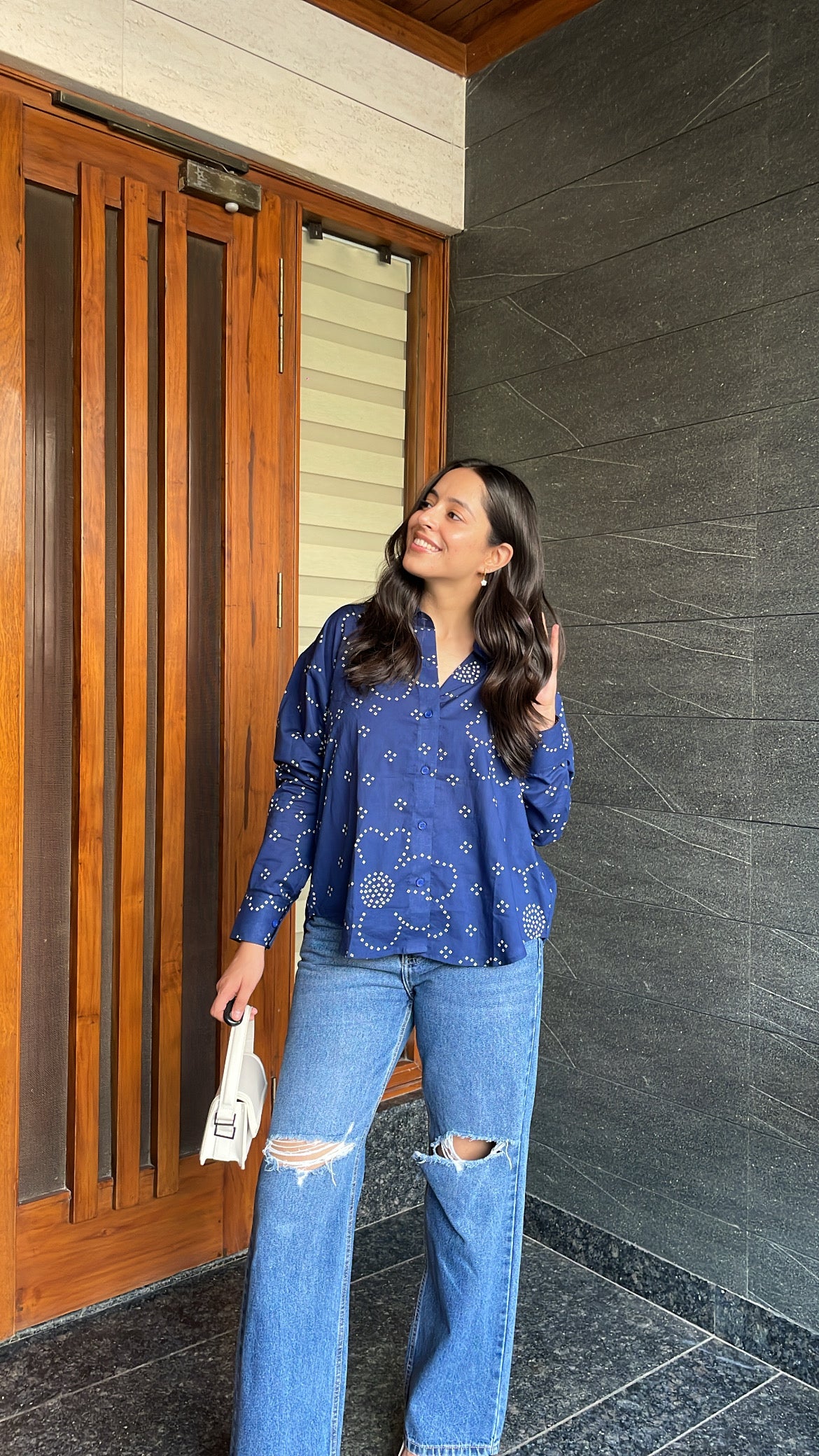Woman in a blue patterned top and jeans standing in front of a wooden door.