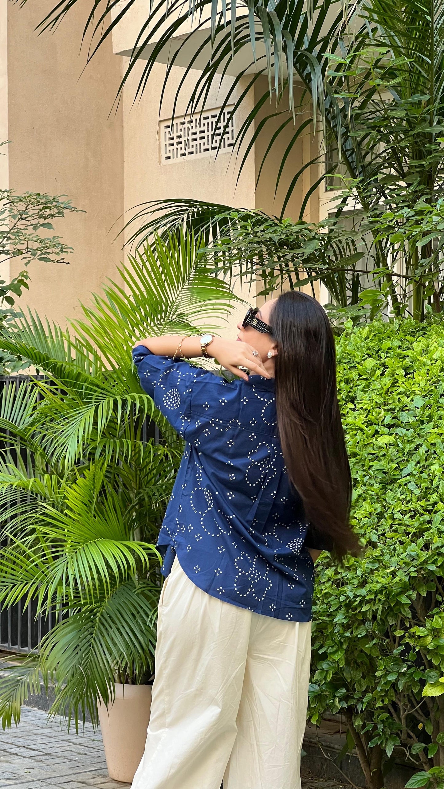 Woman in a blue patterned shirt and white pants standing outdoors with greenery around.