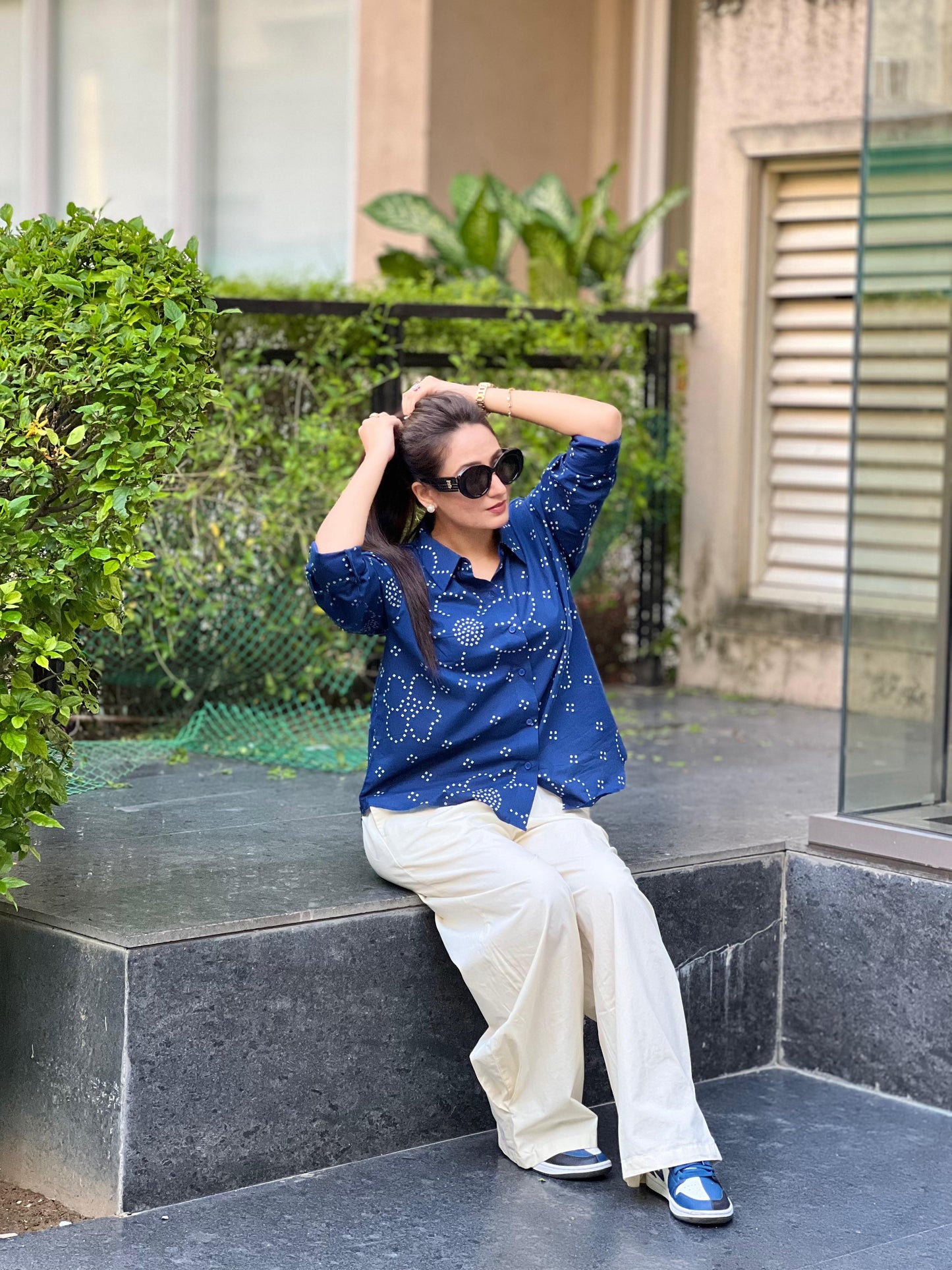 Woman sitting on a stone ledge outdoors, wearing a blue patterned shirt and white pants.