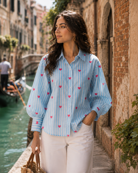 A woman stands on a canal-side walkway wearing a light blue and white striped button-up blouse with small pink hearts, paired with white pants. The background shows a real outdoor environment with brick buildings, water, and a gondola.