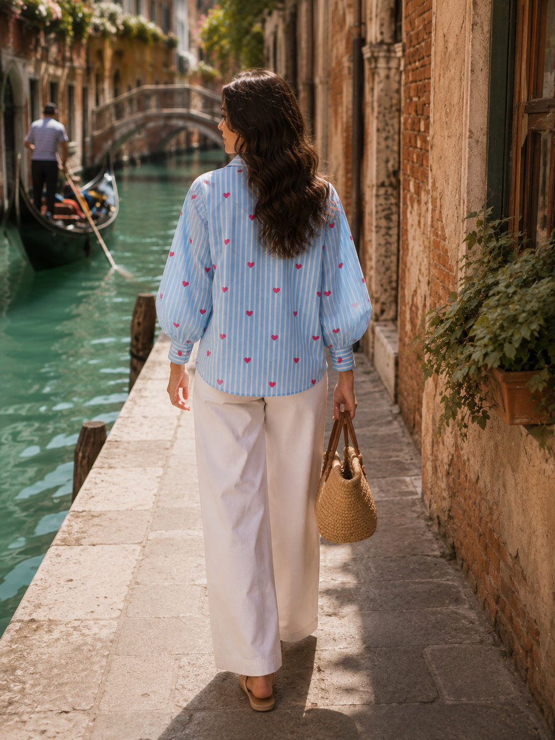 A woman walking along a stone canal-side path in Venice, seen from behind. She is wearing a light blue and white striped blouse with small red embroidered motifs, white wide-leg trousers, and carrying a woven straw handbag.
