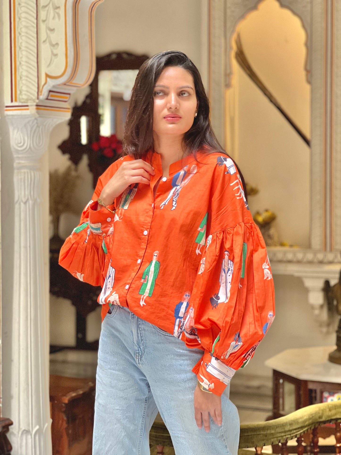Woman wearing an orange patterned blouse and light blue jeans indoors.