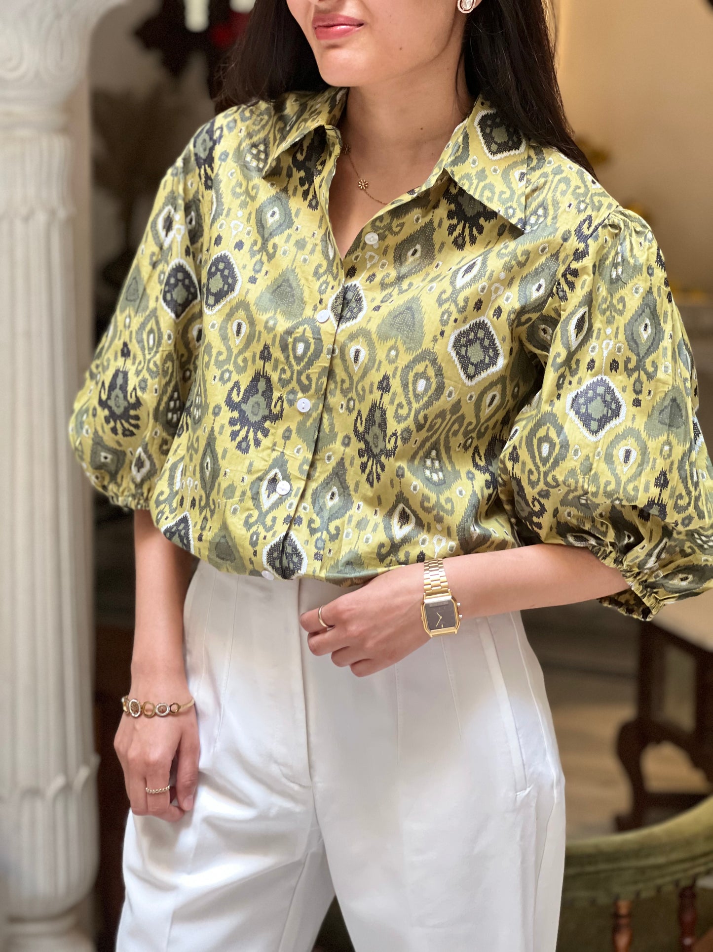 Woman wearing a patterned yellow blouse and white pants indoors.