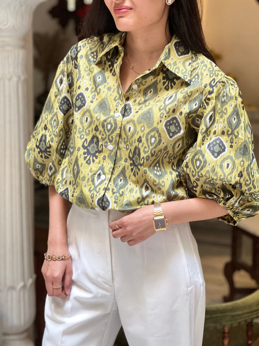 Woman wearing a patterned yellow blouse and white pants indoors.