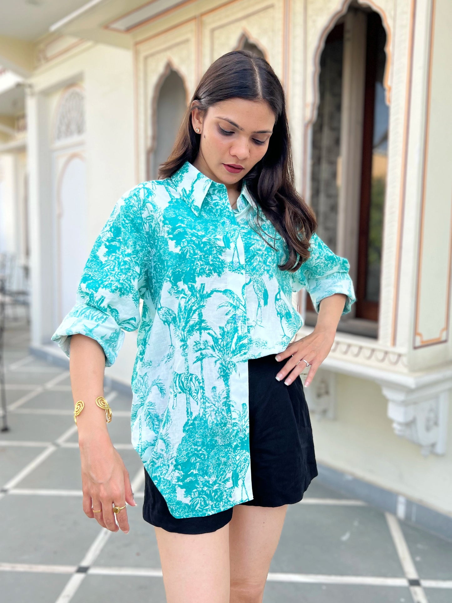 Woman wearing a turquoise patterned shirt and black shorts standing in front of an architectural building.
