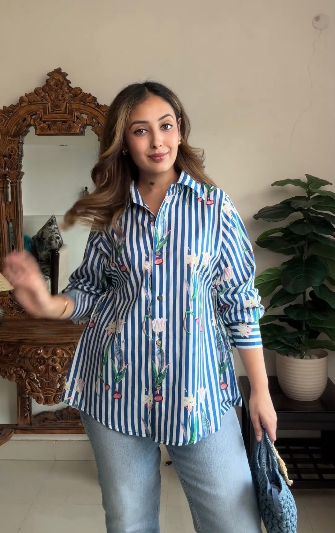 A woman wearing a blue and white striped button-up shirt with a floral print, paired with light blue jeans, standing in an indoor setting with a mirror and a potted plant in the background.