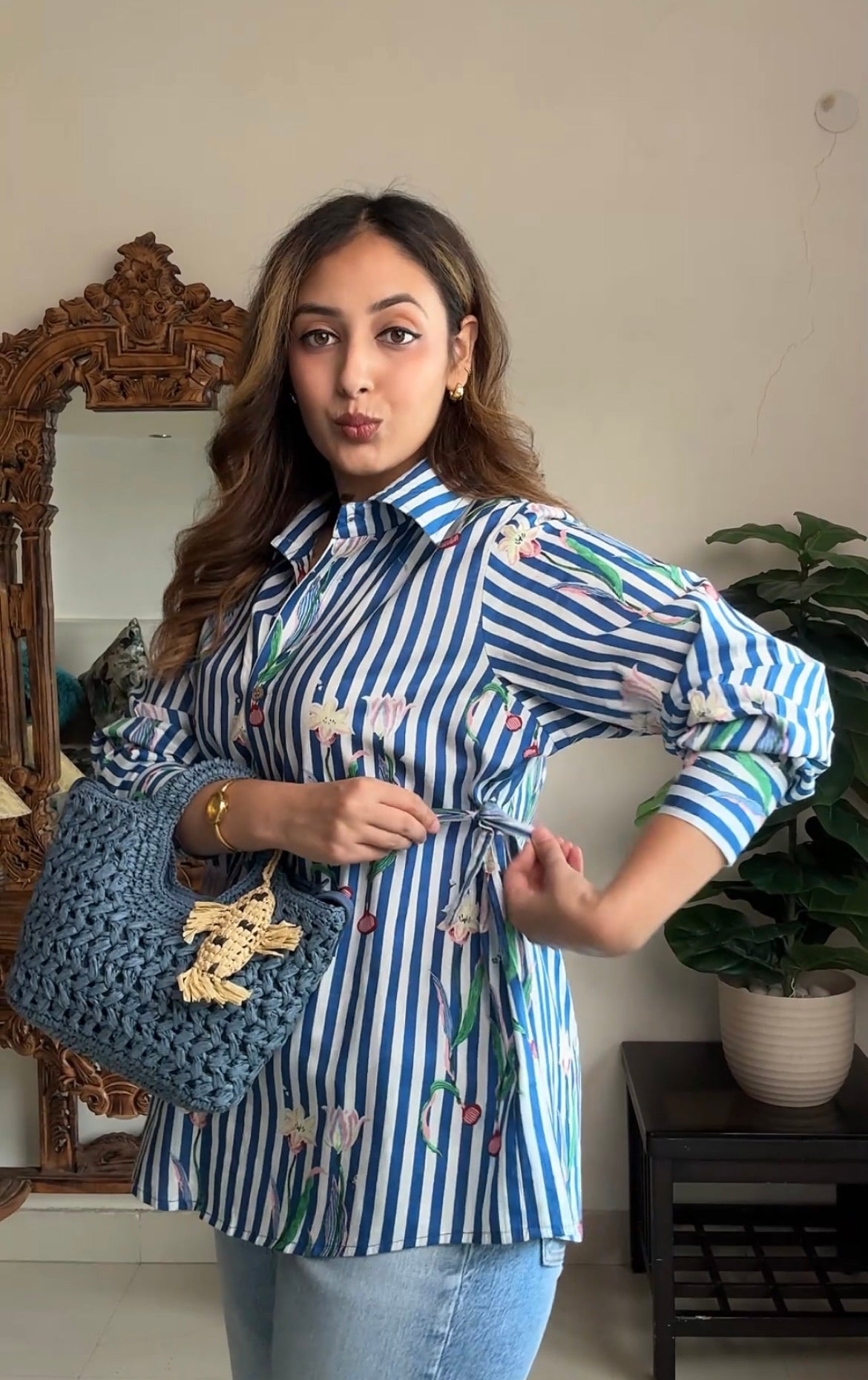 A woman wearing a blue and white striped long-sleeve shirt with a colorful floral and bird pattern, paired with light blue jeans and holding a blue crocheted handbag.