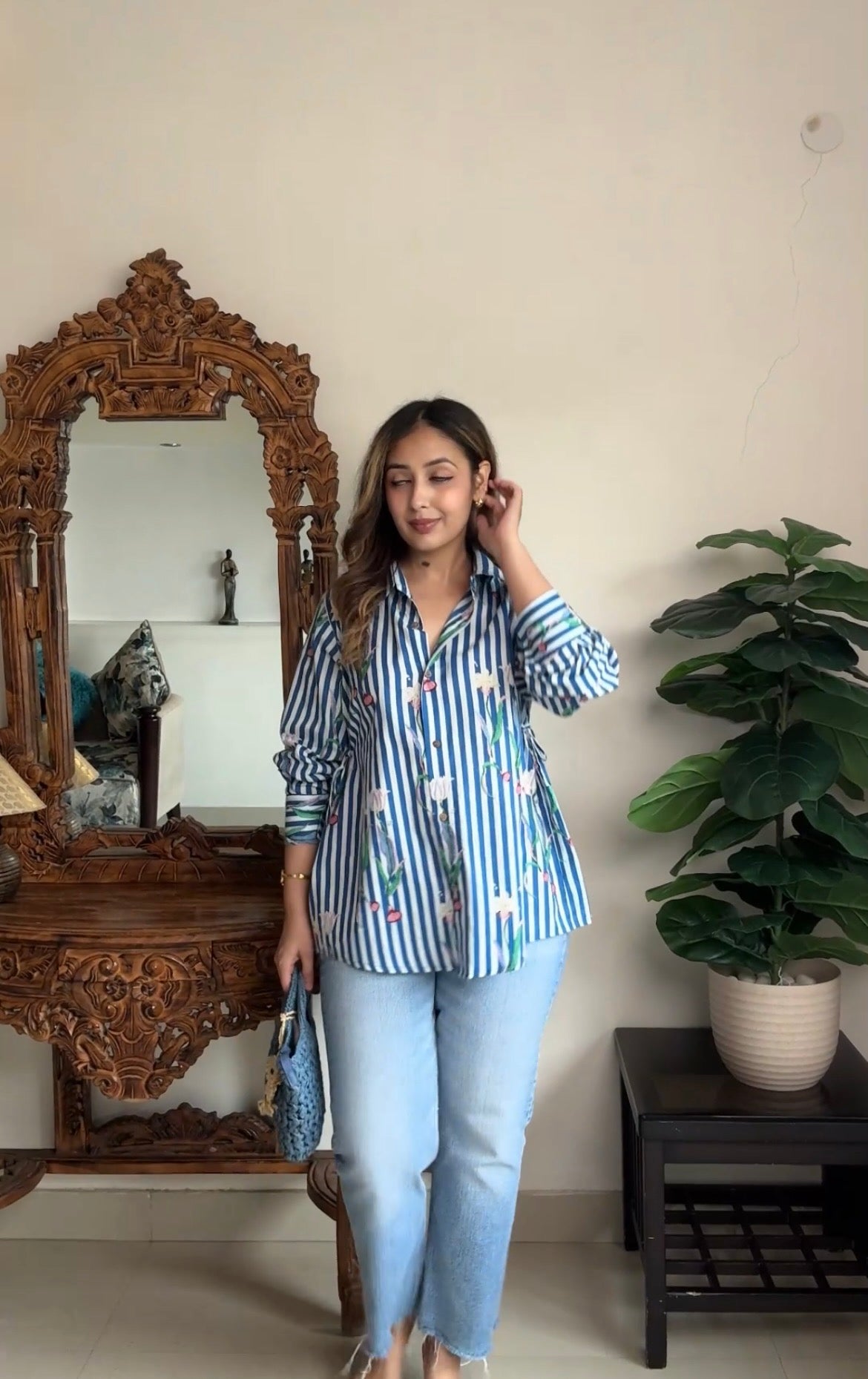 A woman stands indoors wearing a blue and white striped button-up shirt with a small colorful allover print, paired with light blue jeans. She is posed in front of a large ornate wooden mirror and next to a potted green plant.