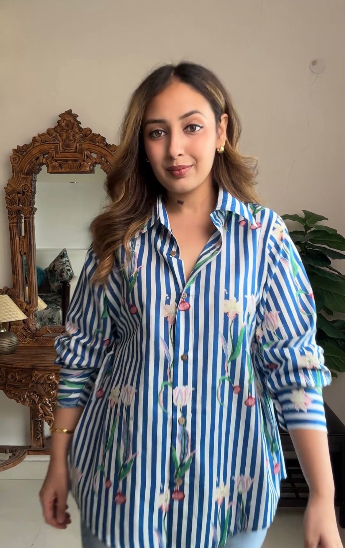 A woman wearing a blue and white vertically striped button-up shirt with a floral tulip print, standing in an indoor setting with a wooden mirror and plant in the background.