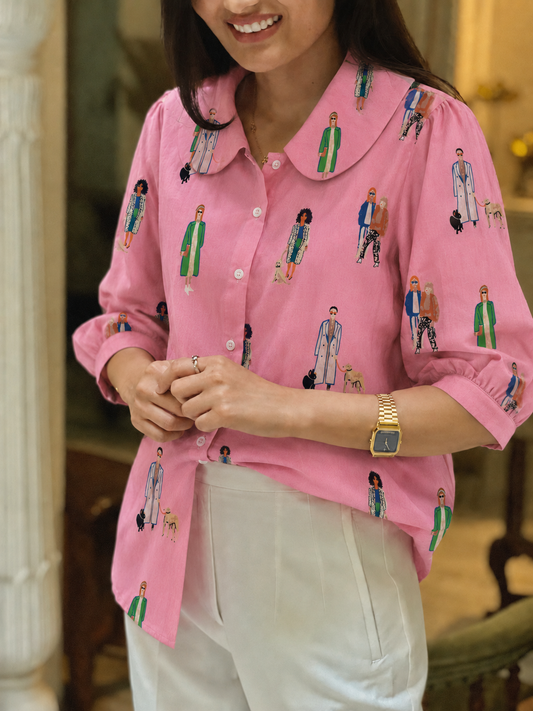 A woman wearing a pink long-sleeve button-up shirt with a Peter Pan collar and an all-over embroidered figure pattern, paired with white trousers in an indoor setting.