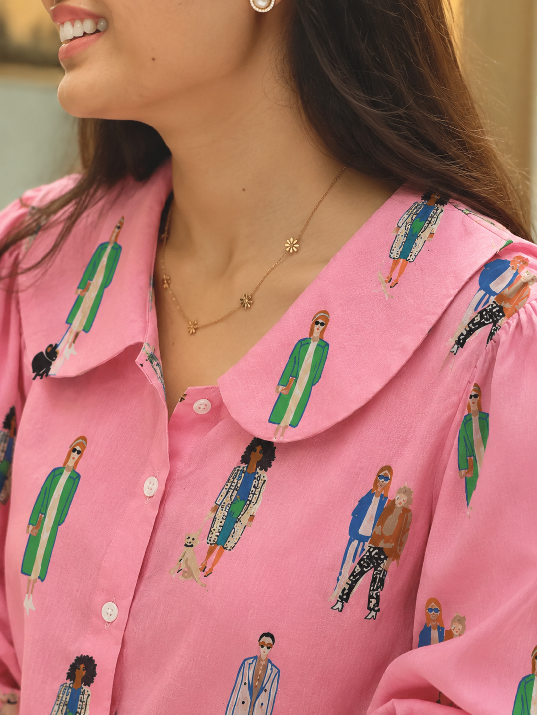 A close-up of a woman wearing a pink button-up shirt with a Peter Pan collar, featuring a colorful all-over print of illustrated fashion figures. She is also wearing a delicate gold necklace.
