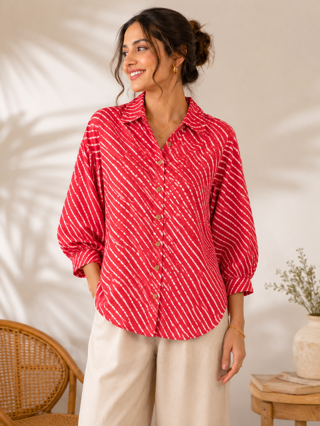A woman wearing a red and white striped button-up shirt with beige wide-leg pants, standing in a softly lit indoor setting with a wicker chair and small table in the background.