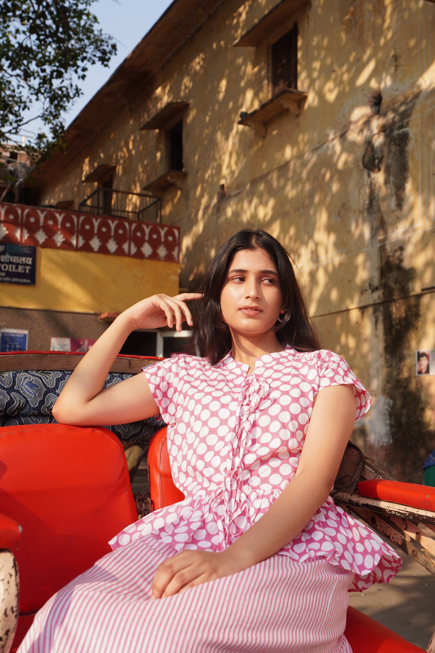 Woman in a pink dress sitting on a red chair outdoors with a building and tree in the background.
