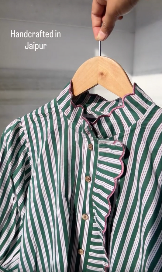 A green and white vertically striped blouse with pink scalloped trim and brown buttons, hanging on a wooden hanger held by a hand. The text 'Handcrafted in Jaipur' is visible on the top left.