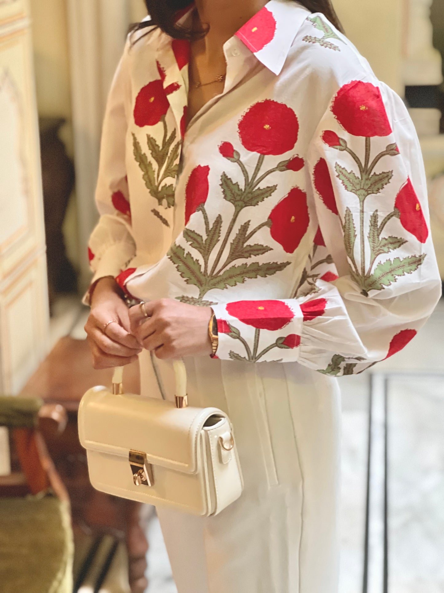 Person wearing a floral blouse with red flowers and beige pants, holding a beige handbag.
