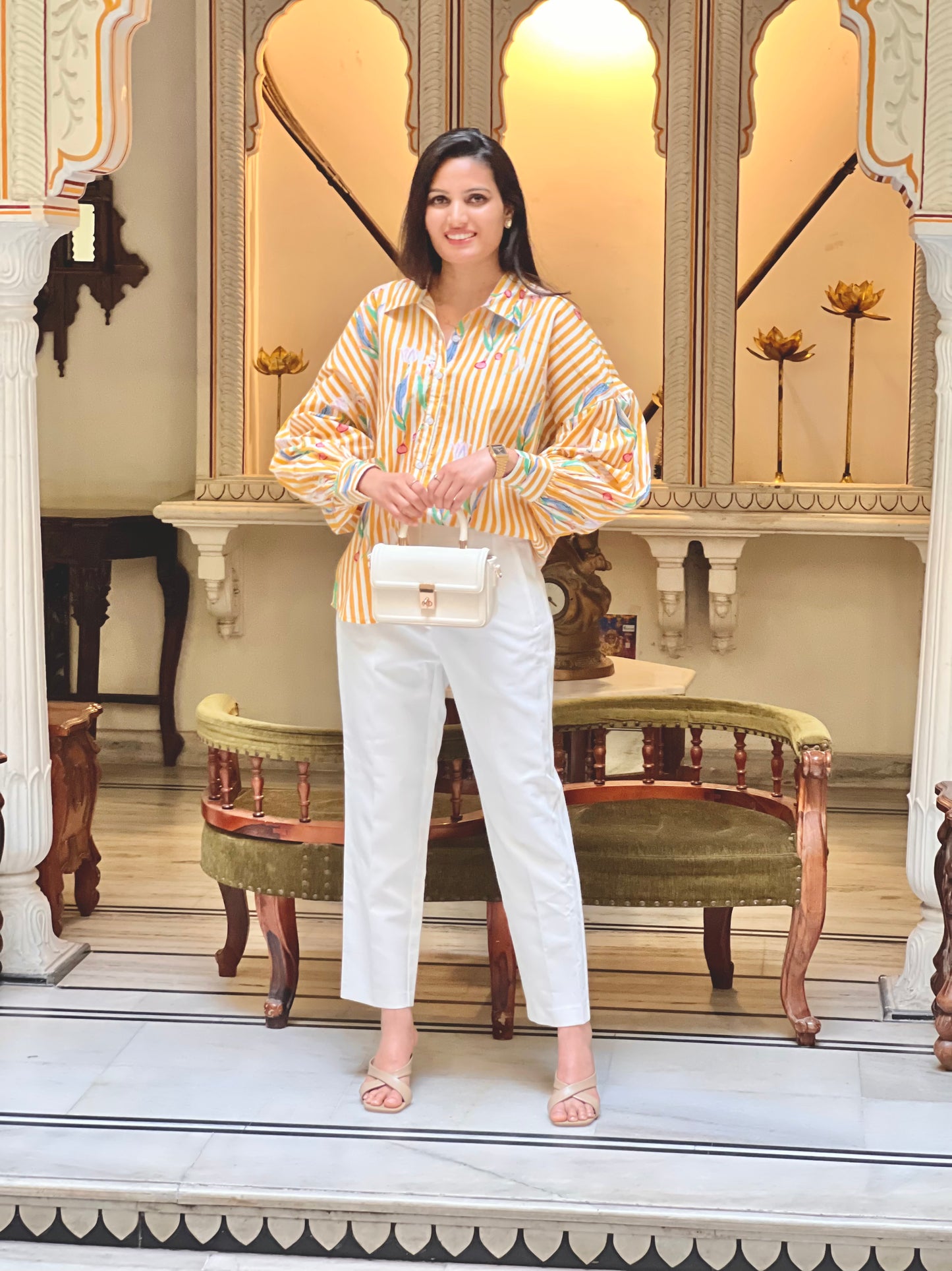 Woman in a patterned top and white pants holding a handbag in an ornate interior setting.