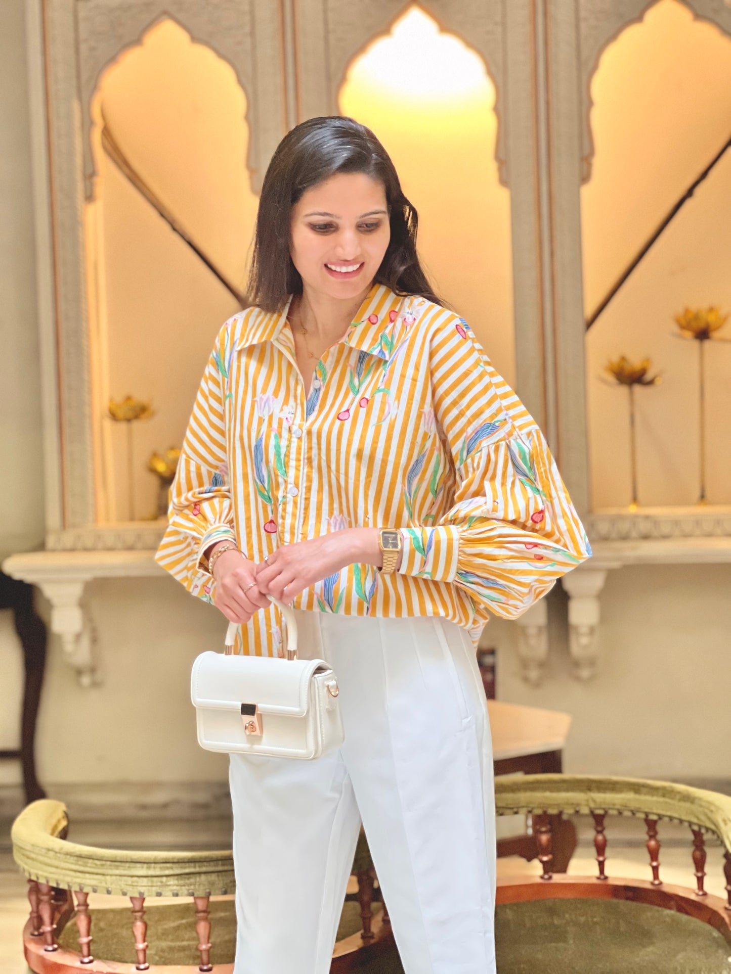 Woman in a yellow striped shirt and white pants standing in an elegant interior setting.