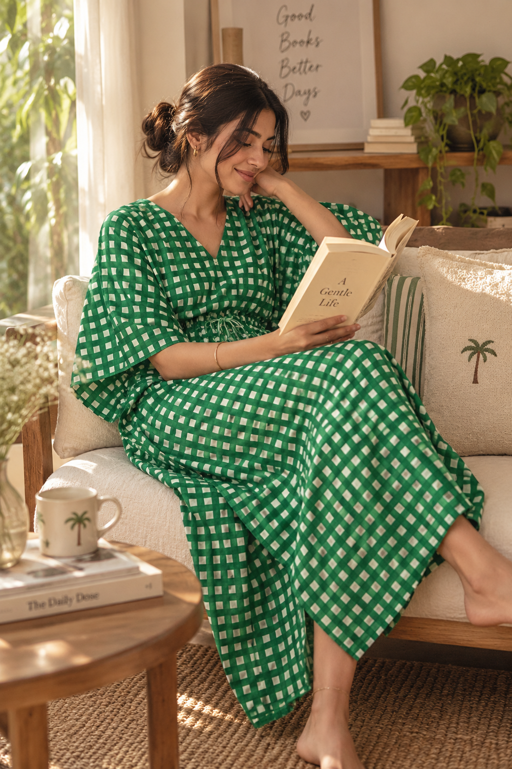 A woman sits on a beige sofa in a sunlit living room, reading a book while wearing a green and white gingham maxi dress. A wooden side table with a mug is in the foreground, and framed wall art and plants are visible in the background.
