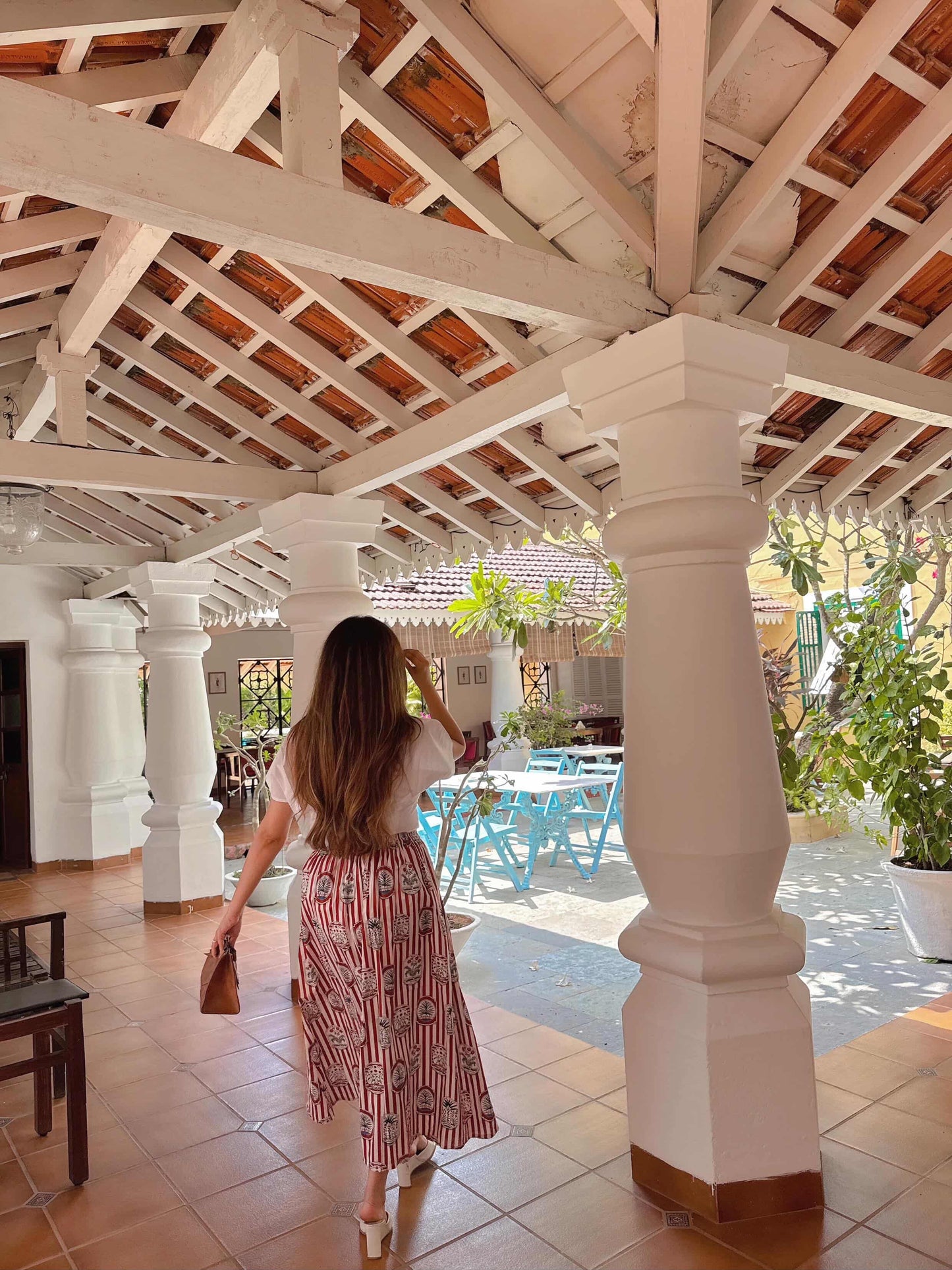 Woman standing in a sunlit indoor area with wooden ceiling and white columns, looking towards a pool.