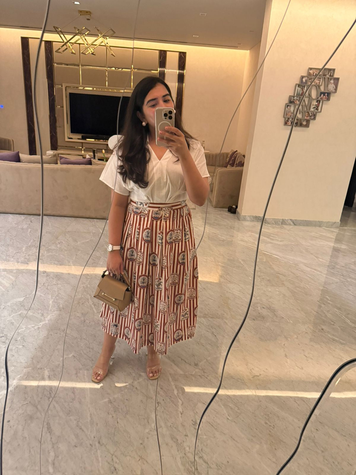 Woman taking a mirror selfie wearing a white blouse and patterned skirt in a modern interior setting.